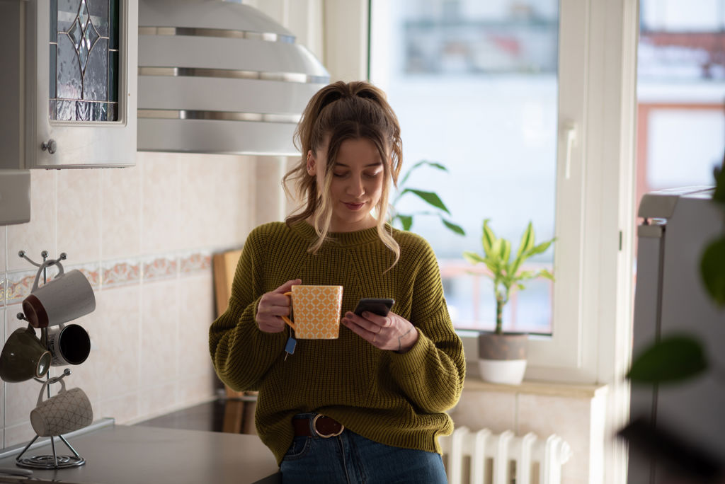 Vrouw op telefoon in de keuken met een mok thee.